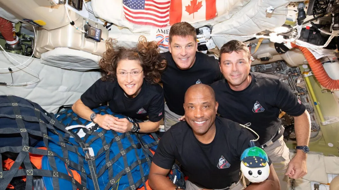 Four astronauts smiling in space station, with US and Canadian flags.