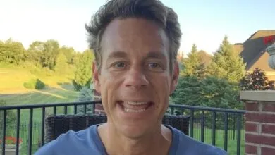 Man smiles outdoors on balcony with trees and house in background.