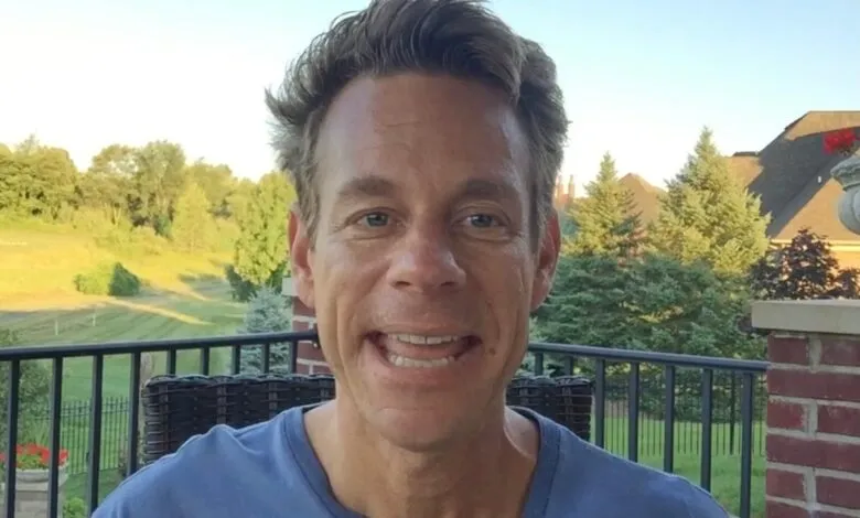 Man smiles outdoors on balcony with trees and house in background.
