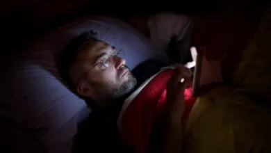 Man with glasses lies in bed, illuminated by phone screen.