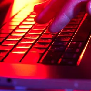 Close-up of a hand typing on a laptop keyboard, illuminated with red and yellow light.