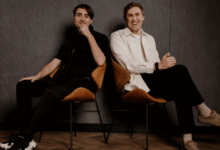 Two men sit in chairs against a dark wall, one smiling, the other thoughtful.