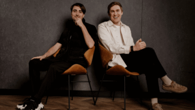 Two men sit in chairs against a dark wall, one smiling, the other thoughtful.