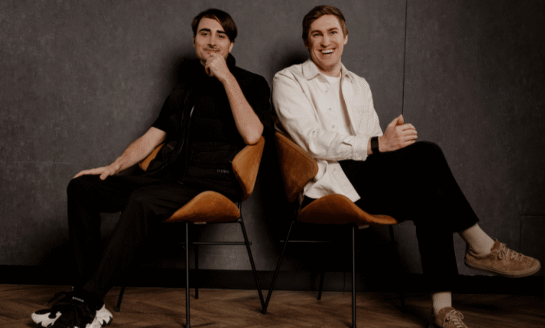 Two men sit in chairs against a dark wall, one smiling, the other thoughtful.