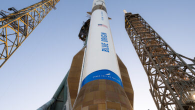 Blue Origin rocket on launchpad, surrounded by support towers, against a clear sky.