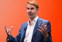 John Doerr gestures while speaking on stage with orange backdrop.