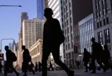 Silhouetted pedestrians cross a busy Chicago street with tall buildings in the background.