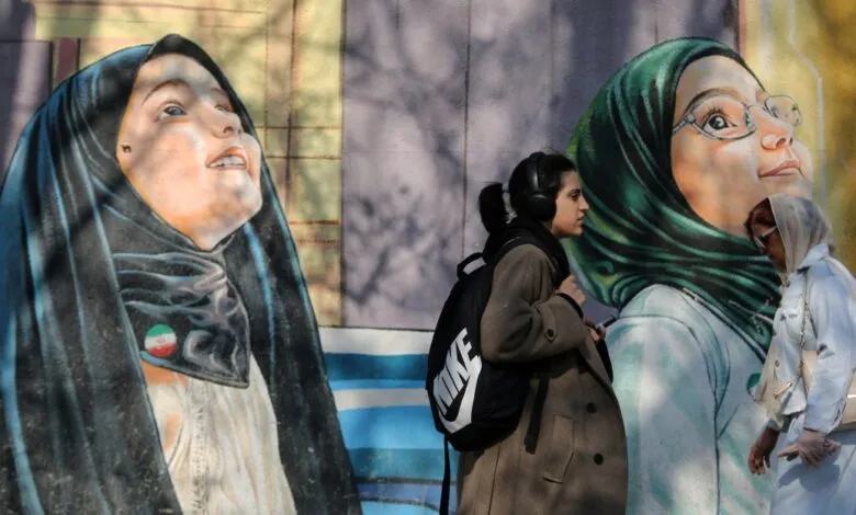 Two women walk past murals of women in hijabs on a sunny day.