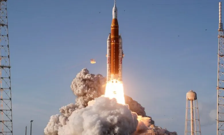 NASA's Artemis rocket launches, surrounded by smoke and flames against a blue sky.