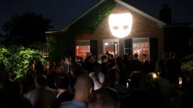 Outdoor party at night with crowd and projected face on brick building.