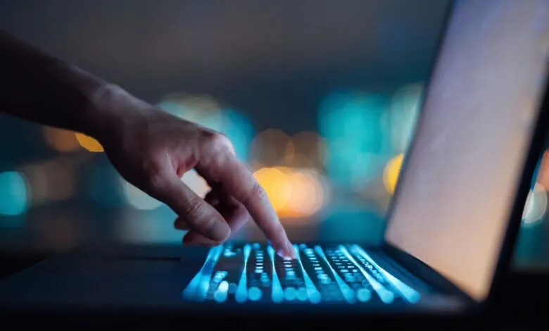Hand types on a laptop keyboard with blue backlighting in a dark room.