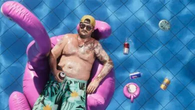 Man relaxes in pool on flamingo float with drinks and blue tiles.