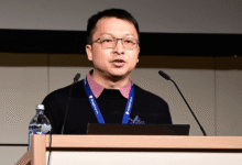 Man with glasses presents at podium with microphones and water bottle.