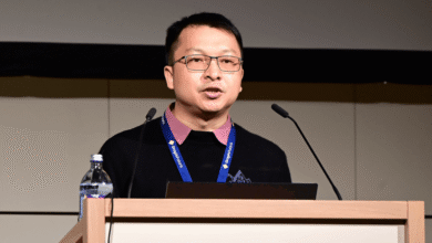 Man with glasses presents at podium with microphones and water bottle.