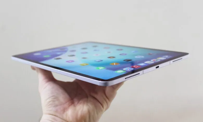Hand holding a thin, light purple iPad Air showing the home screen.