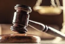 Wooden gavel rests on block with scales of justice in background.