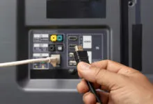 Hand plugging black HDMI cable into the back of a gray television.