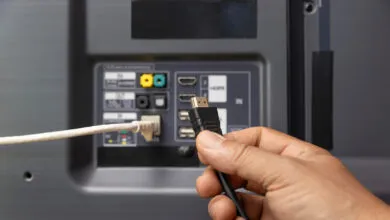 Hand plugging black HDMI cable into the back of a gray television.