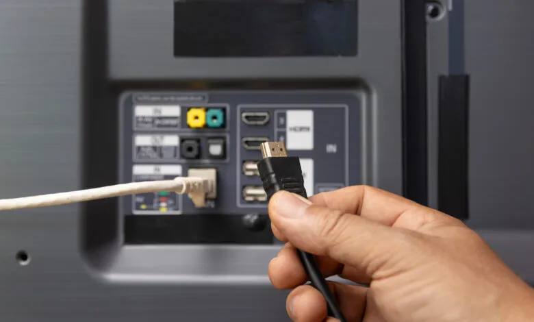 Hand plugging black HDMI cable into the back of a gray television.