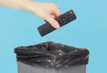 Hand dropping black remote control into trash can with blue background.