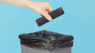 Hand dropping black remote control into trash can with blue background.