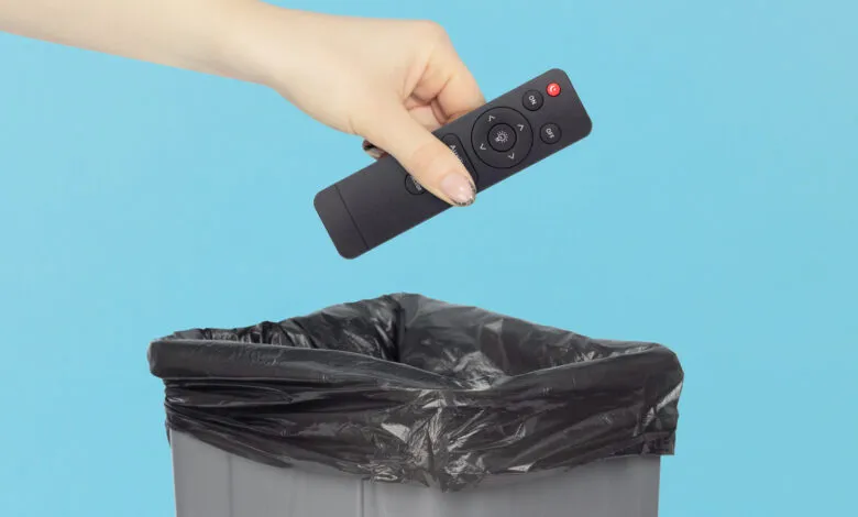 Hand dropping black remote control into trash can with blue background.