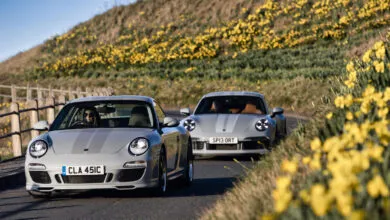 Two gray Porsche sports cars driving on a road lined with yellow flowers.