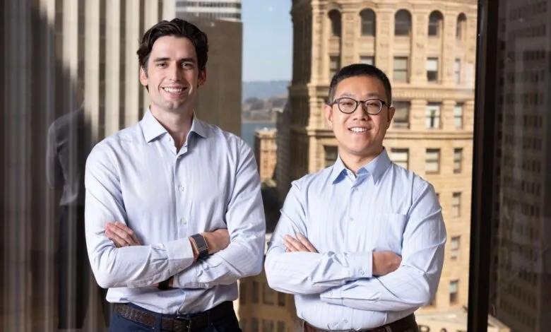 Two smiling men stand with arms crossed in front of city skyline.