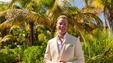 Man in cream suit smiles against tropical palm tree backdrop.