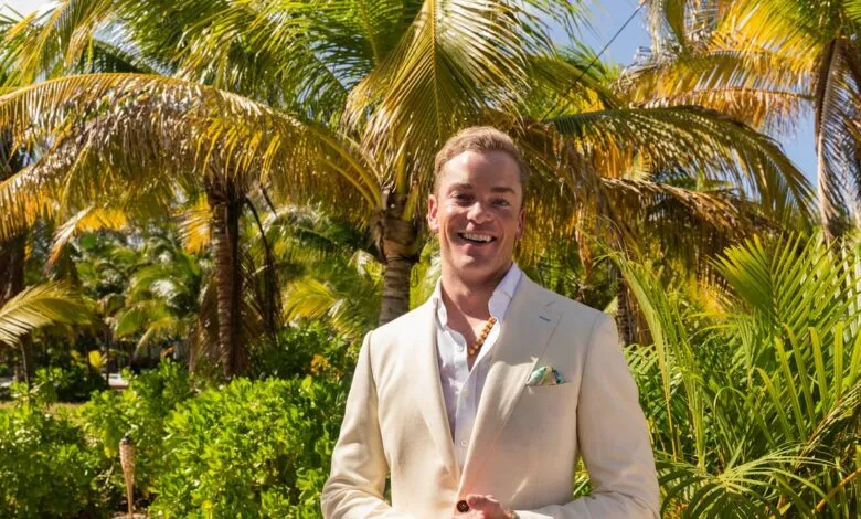 Man in cream suit smiles against tropical palm tree backdrop.
