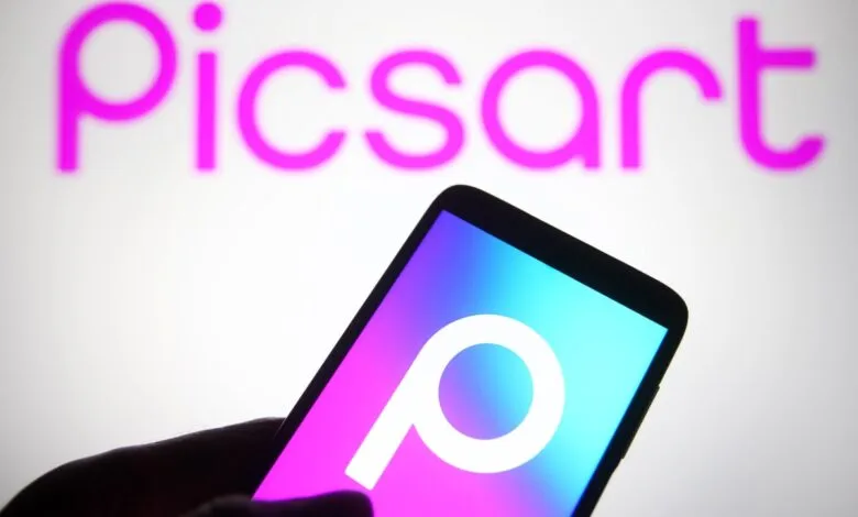 Hand holding phone displaying Picsart logo against blurred Picsart background.