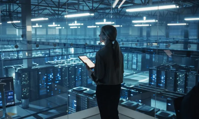 Woman with tablet overlooking a large server room through glass.