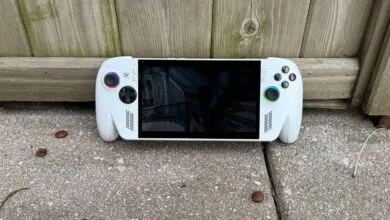 White handheld gaming PC displaying a game, resting against a wooden fence.