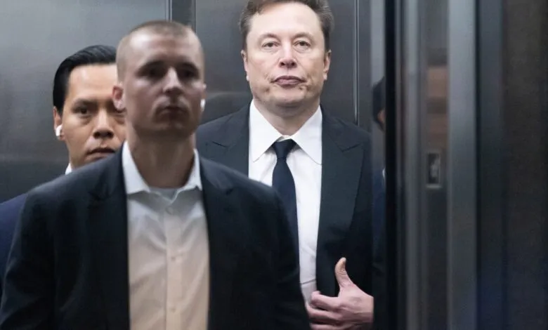 Elon Musk in suit with two men in elevator, serious expression.
