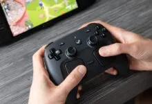 Hands hold black controller, Steam Deck visible in background.