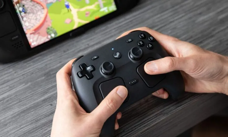 Hands hold black controller, Steam Deck visible in background.