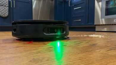 Black robot vacuum cleaner with green light on wood floor near spilled rice.