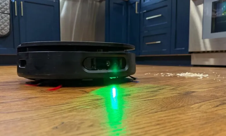 Black robot vacuum cleaner with green light on wood floor near spilled rice.