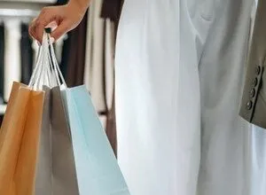 Person holding shopping bags in a clothing store, wearing white pants and a jacket.