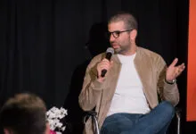 Man with glasses speaks into microphone, gesturing with his hands.