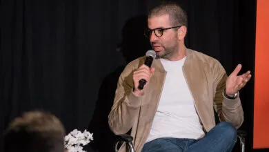 Man with glasses speaks into microphone, gesturing with his hands.