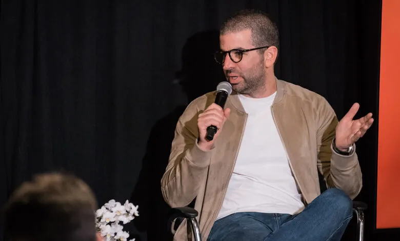 Man with glasses speaks into microphone, gesturing with his hands.