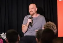 Bald man with beard speaking into microphone on stage.