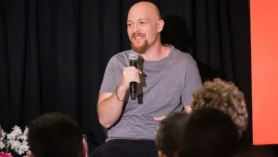 Bald man with beard speaking into microphone on stage.