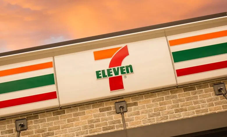 7-Eleven store sign with orange, green, and red stripes against a sunset sky.