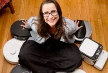 Woman smiling surrounded by various robot vacuum cleaners on wood floor.