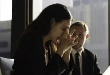 Woman with head in hands sits at desk, man in suit looks on with concern.