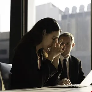 Woman with head in hands sits at desk, man in suit looks on with concern.