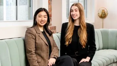 Two young women in business attire sit on a light green couch, smiling at the camera.