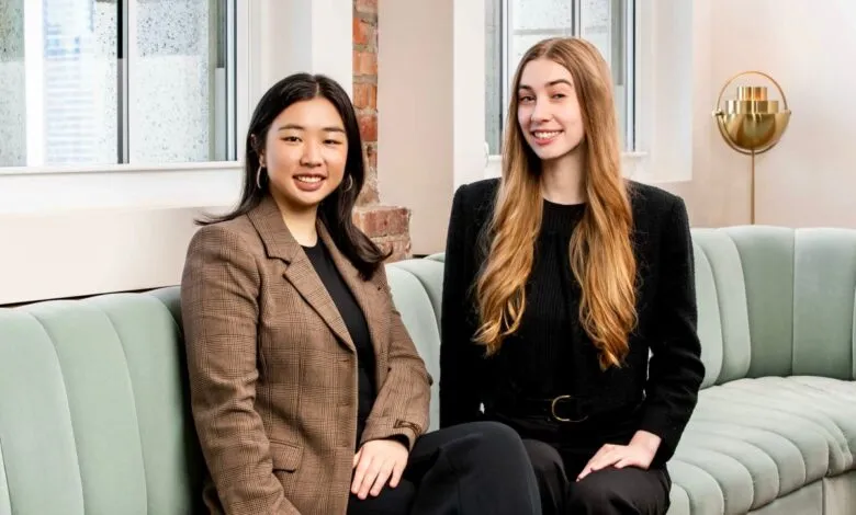 Two young women in business attire sit on a light green couch, smiling at the camera.